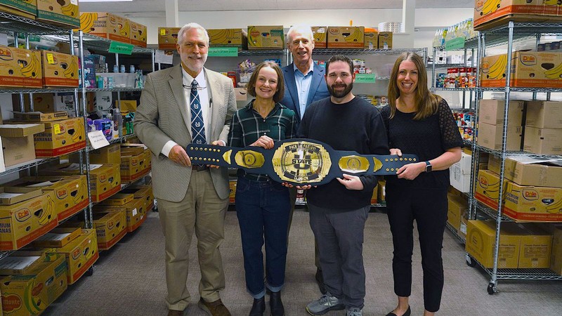 A group of people holding a wrestling-style trophy belt in a food pantry.