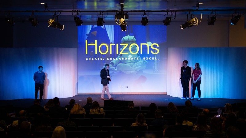 People on stage at an event. Sign text reads: "Horizons. Create. Collaborate. Excel."