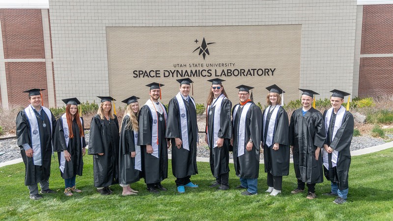 People in graduation robes standing in front of a Space Dynamics Laboratory building.