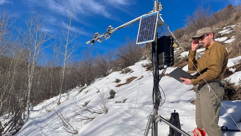 A researcher using a laptop to check a weather station on a snowy hillside.