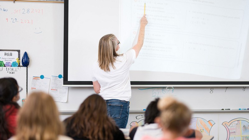 A teacher pointing to a math problem on a projector screen.