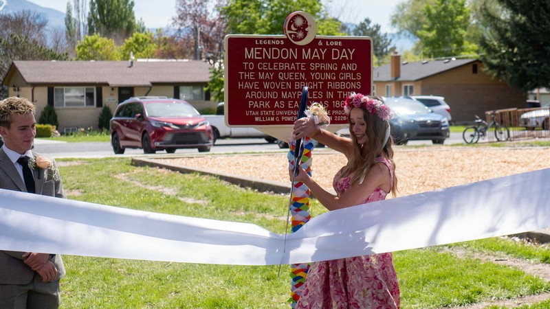 A woman in a dress and traditional May Day flower crown cuts a ribbon for a historical marker.