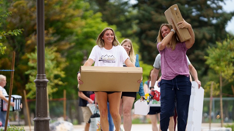 Two women carry boxes as they move things into a dorm room