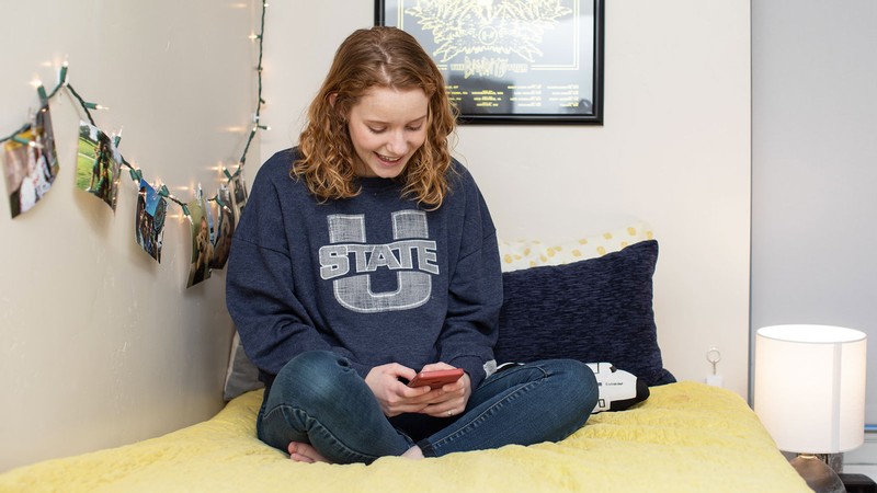 A girl wearing a U State sweatshirt texts as she sits on a bed in a dorm.