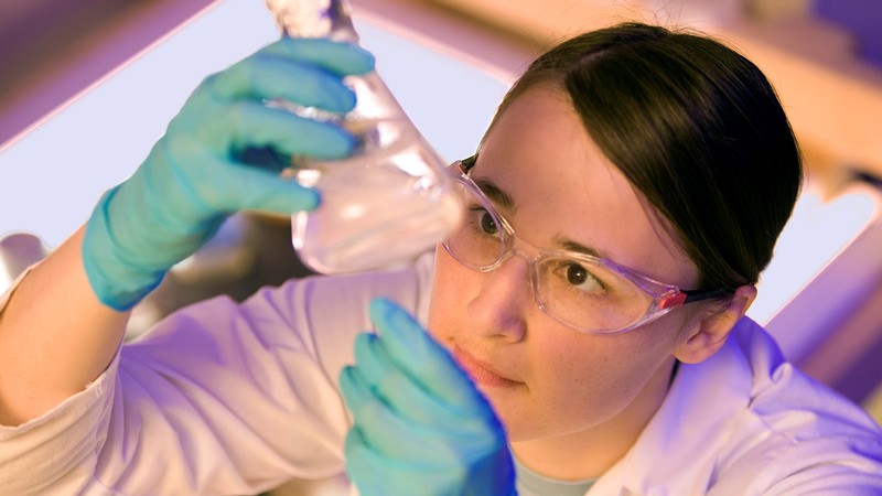 A person wearing safety glasses looks up at liquid in a flask they hold up to the light.