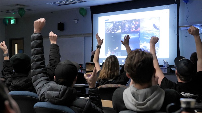 Students cheer while watching a video feed on a projector screen in a dark classroom.