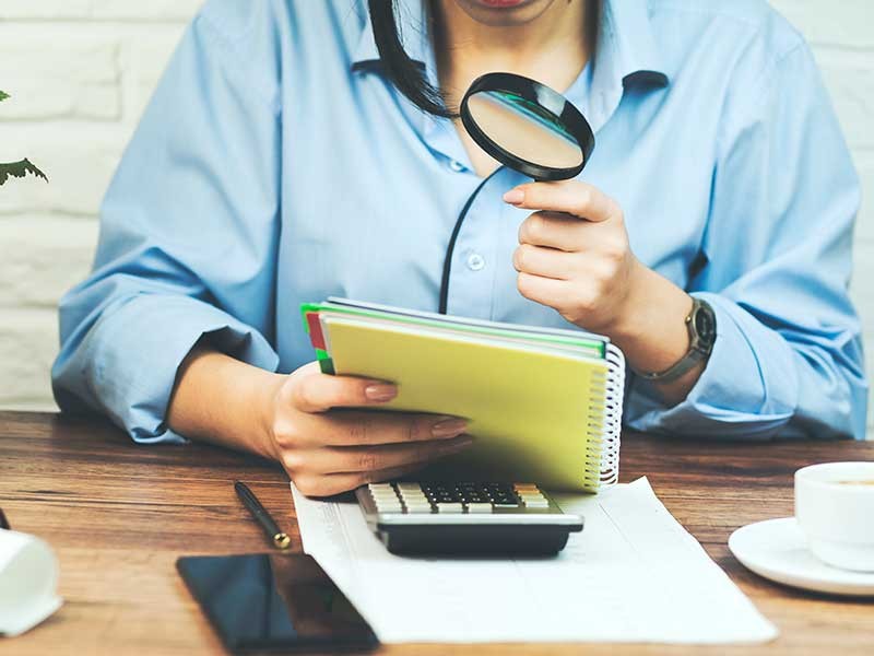 person with a magnifying glass looking over a notebook.