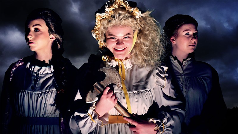 Three actors in period dresses pose for a photo. The character in the middle is holding a rusty axe.