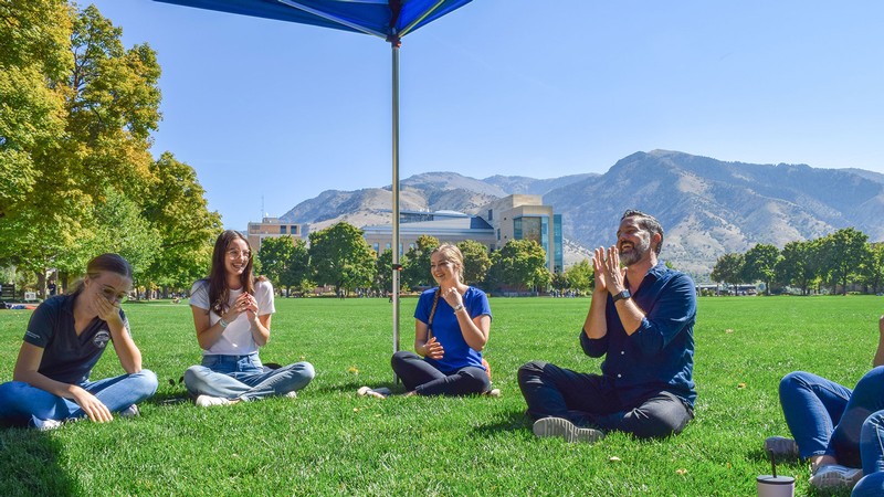 People laugh as they sit on the grass of the USU Quad.