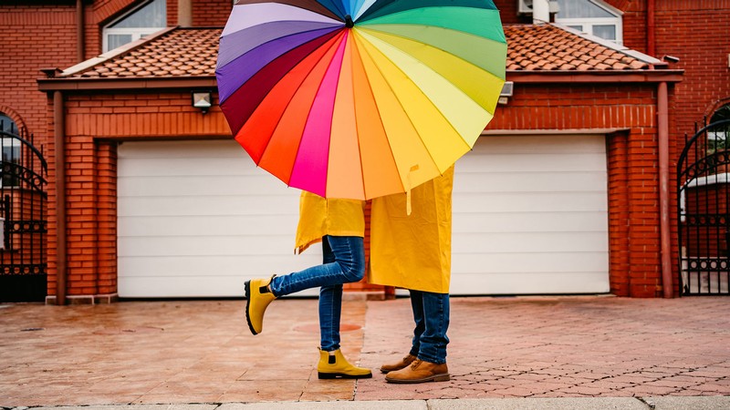 Two people kiss behind a multicolored umbrella.