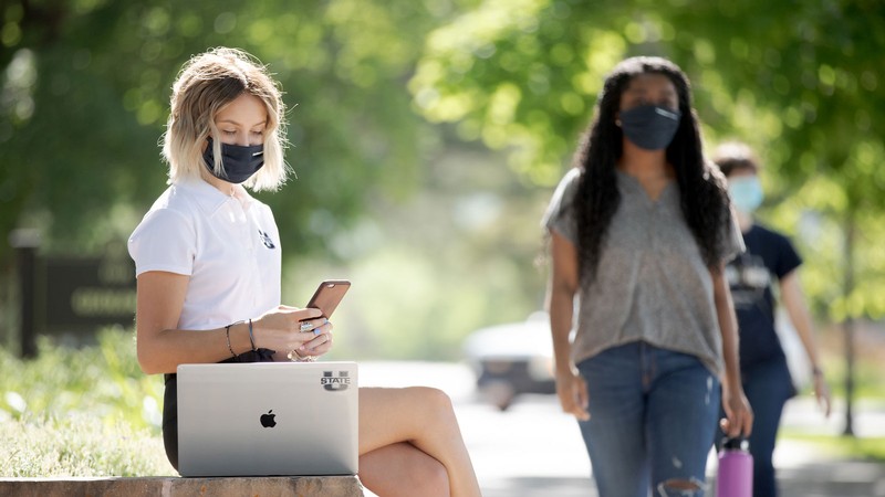 Student on a laptop and wearing a mask with students walking in the background, also wearing masks.