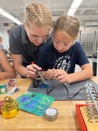 Two girls solder a project in a lab classroom.