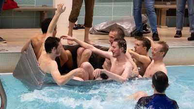 Six students nearly capsizing a boat made of cardboard in a swimming pool.