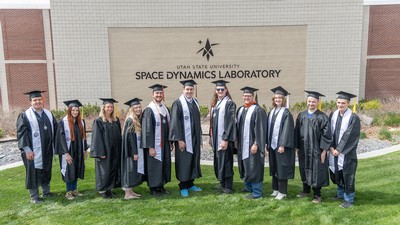 People in graduation robes standing in front of a Space Dynamics Laboratory building.
