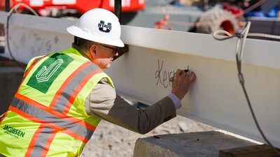 A man in a construction hat and vest signs a steel beam with a marker.