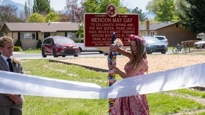 A woman in a dress and traditional May Day flower crown cuts a ribbon for a historical marker.