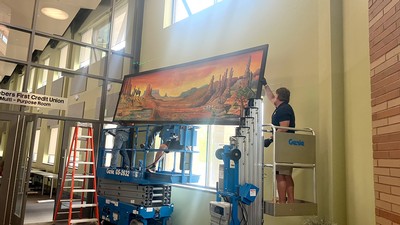 Three people on ladders install a painting of a Southern Utah landscape.