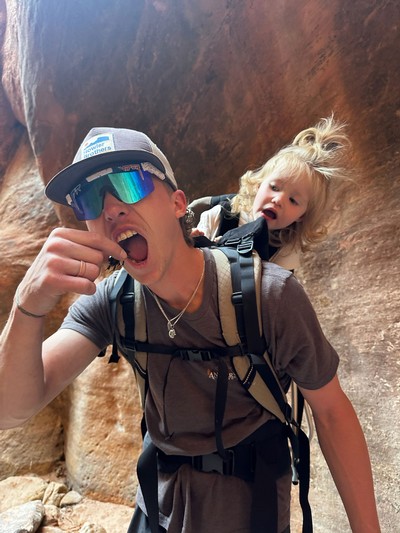 A man appearing to eat a beetle as a worried toddler watches from a child carrier backpack.