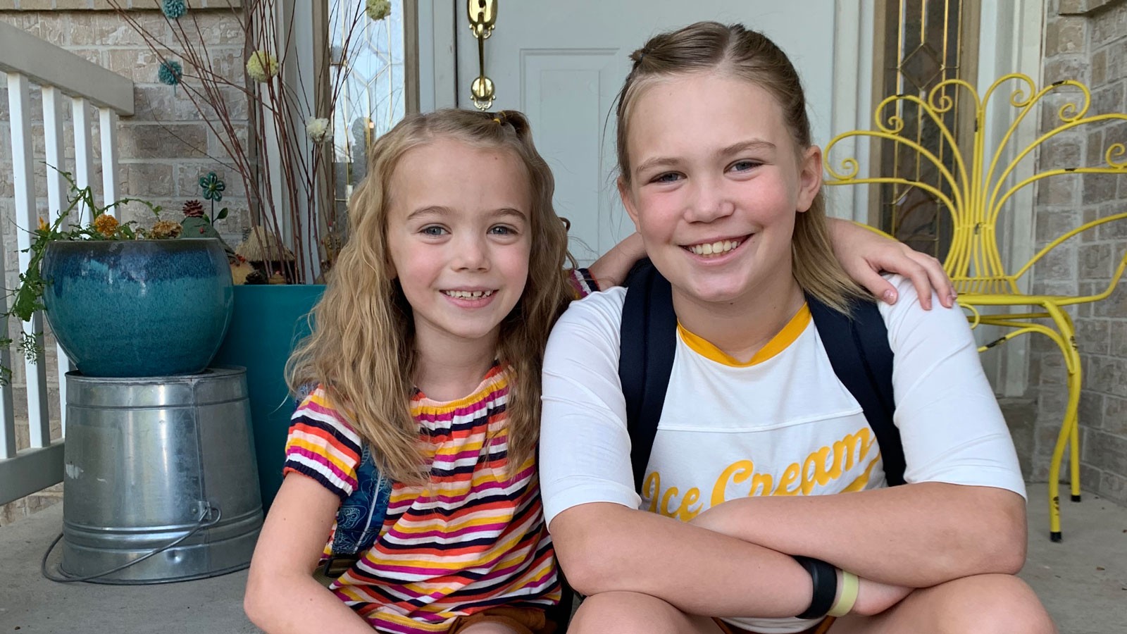 Two school-age children wearing backpacks sit on a porch.