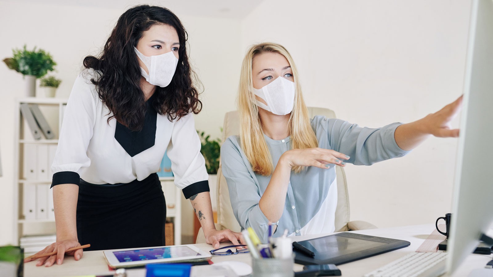 Two people wear masks while working at a computer.