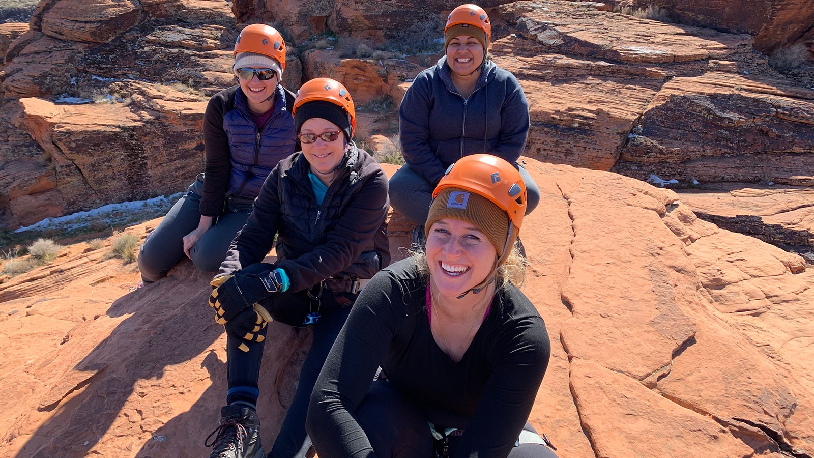 Four women with rock climbing equipment sit atop a rock formation.