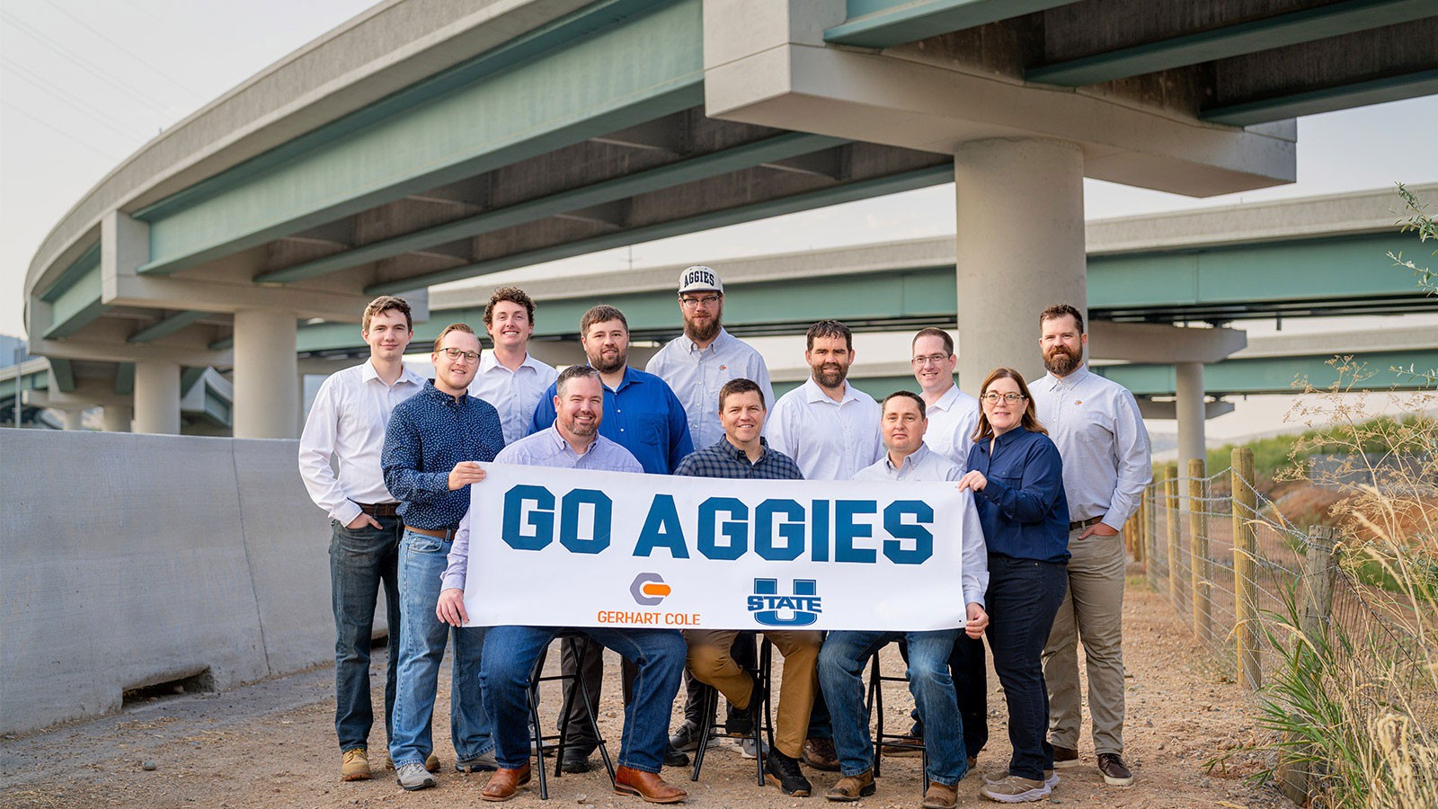 Engineers posing with a banner that says "Go Aggies" near a new highway interchange.