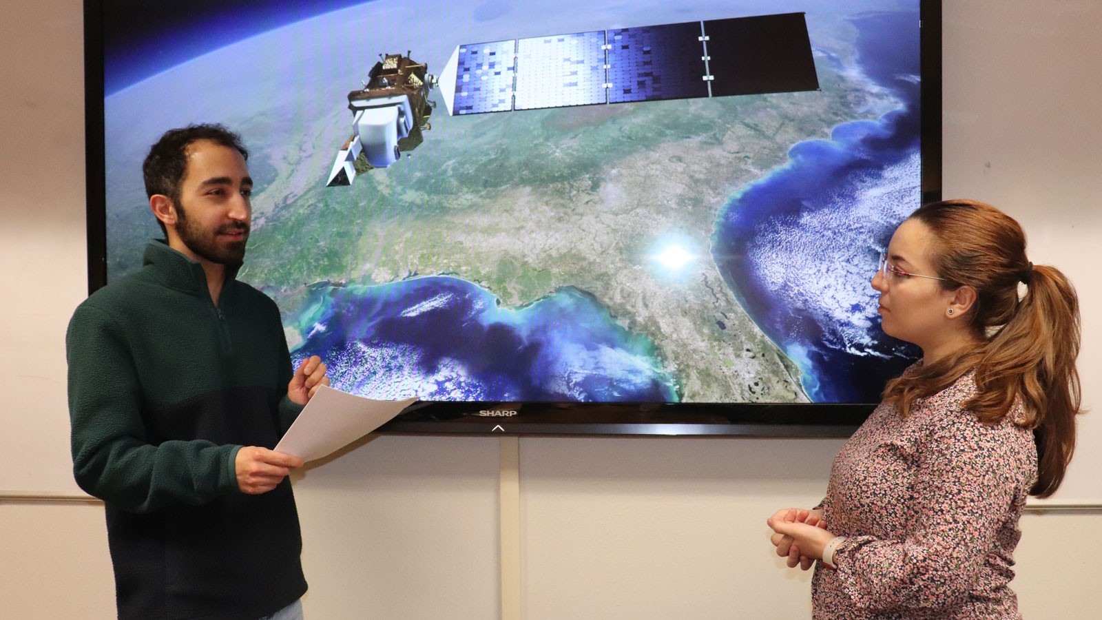 Pouya Hosseinzadeh and Soukaina Filali Boubrahimi stand in front of a TV displaying the image of a satellite. 