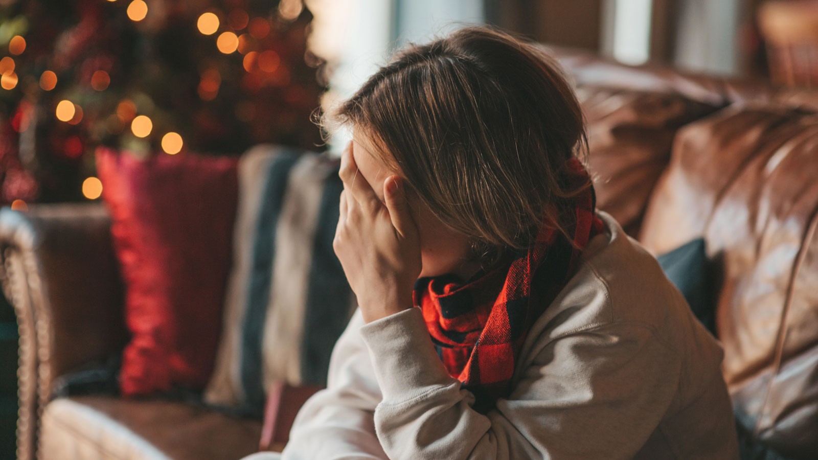 A person with a hand over their face in front of a Christmas tree.