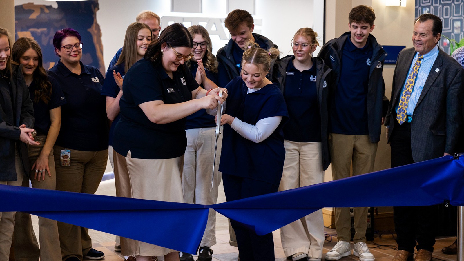 A pair of women hold a giant pair of scissors while cutting a ribbon.