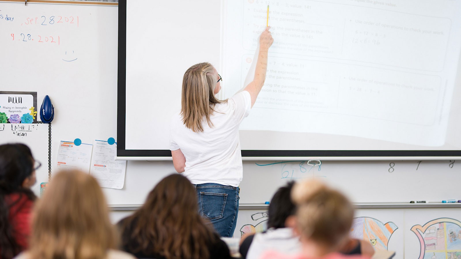 A teacher pointing to a math problem on a projector screen.