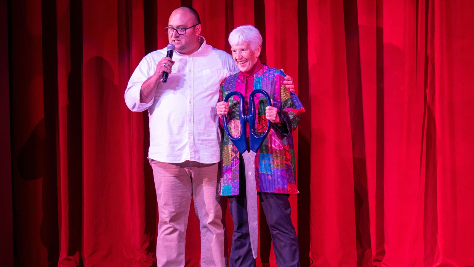 Mary Heers holding a giant pair of scissors on a theater stage with Richie Call.
