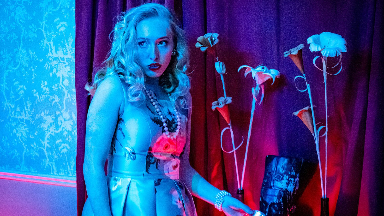 An actor wearing pearls and a dress standing next to flower props.