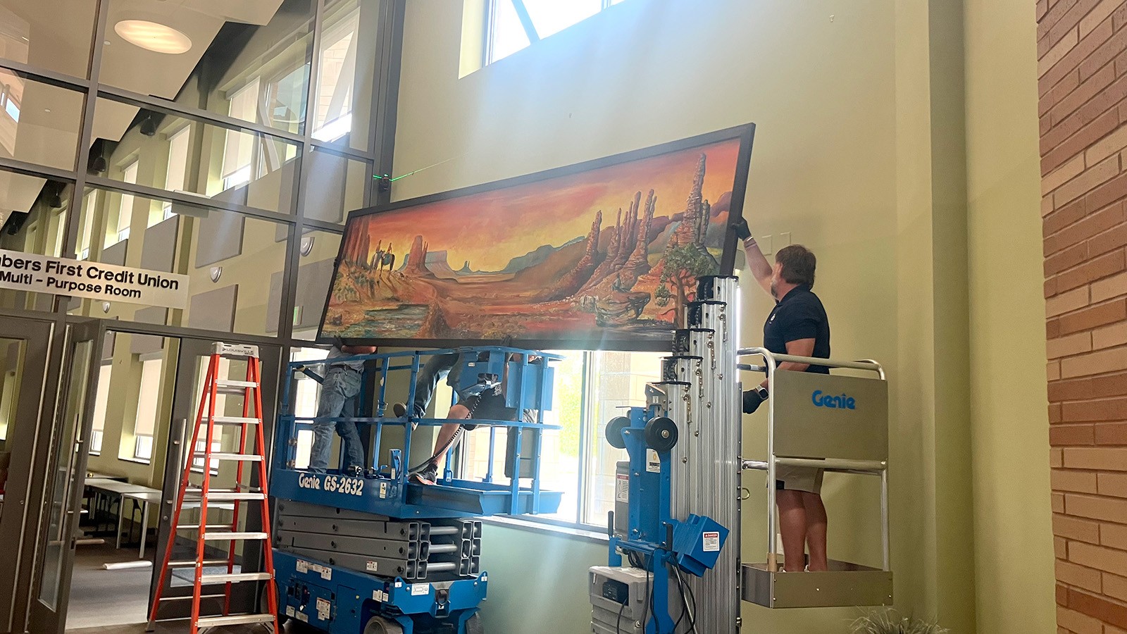 Three people on ladders install a painting of a Southern Utah landscape.