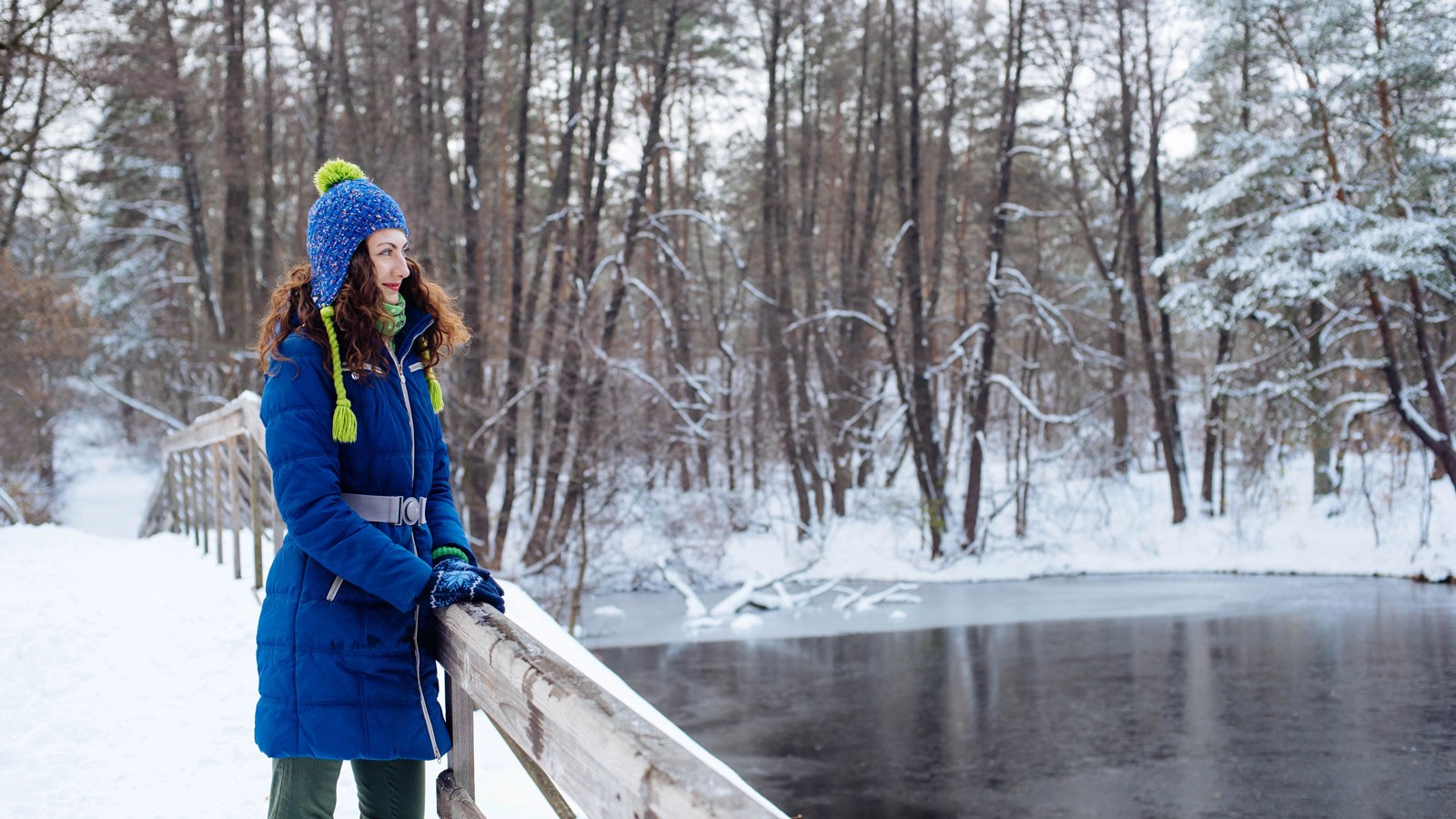 A smiling person in winter clothing standing on a snowy bridge over an icy river.