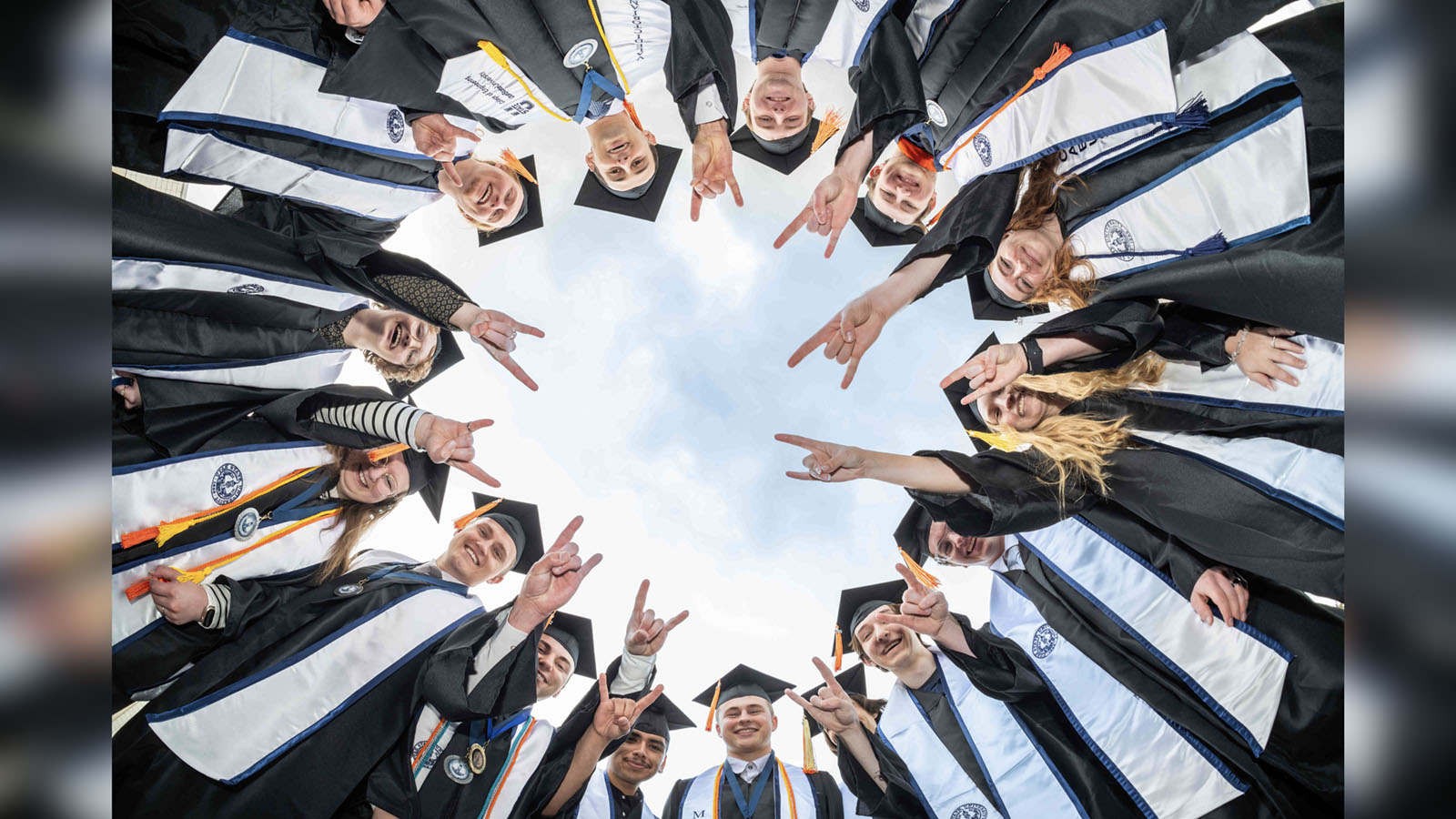 Students posing in a circle and wearing graduation robes make bull horn hand signs.