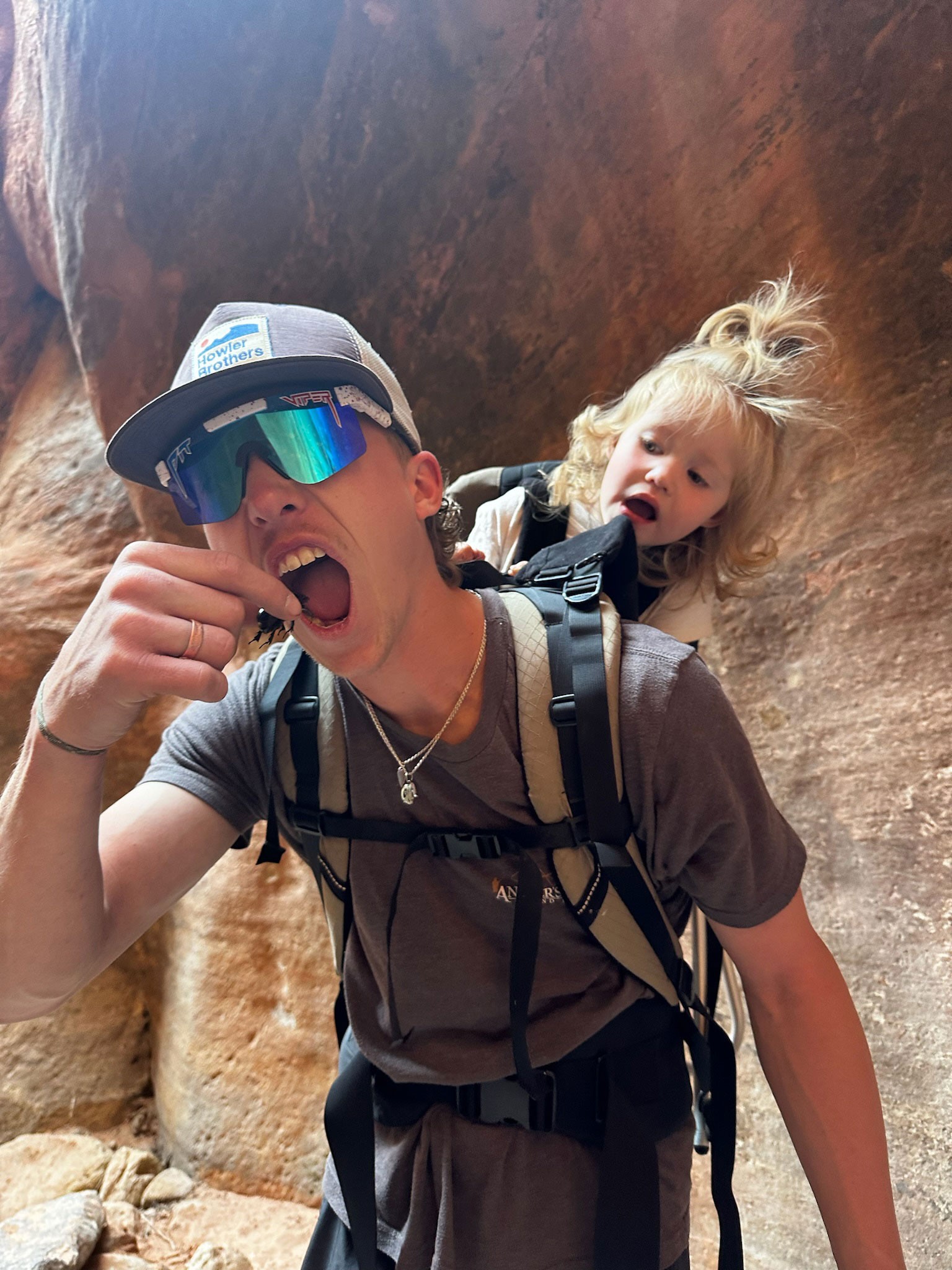 A man appearing to eat a beetle as a worried toddler watches from a child carrier backpack.