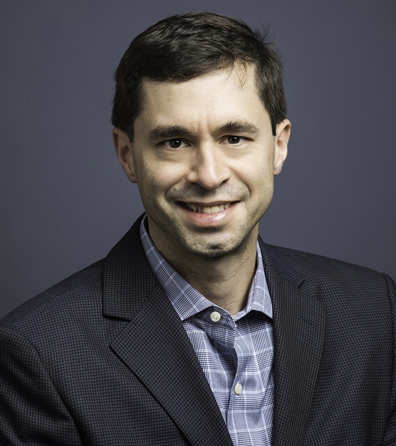 A man in a black suit and blue shirt poses for a headshot.