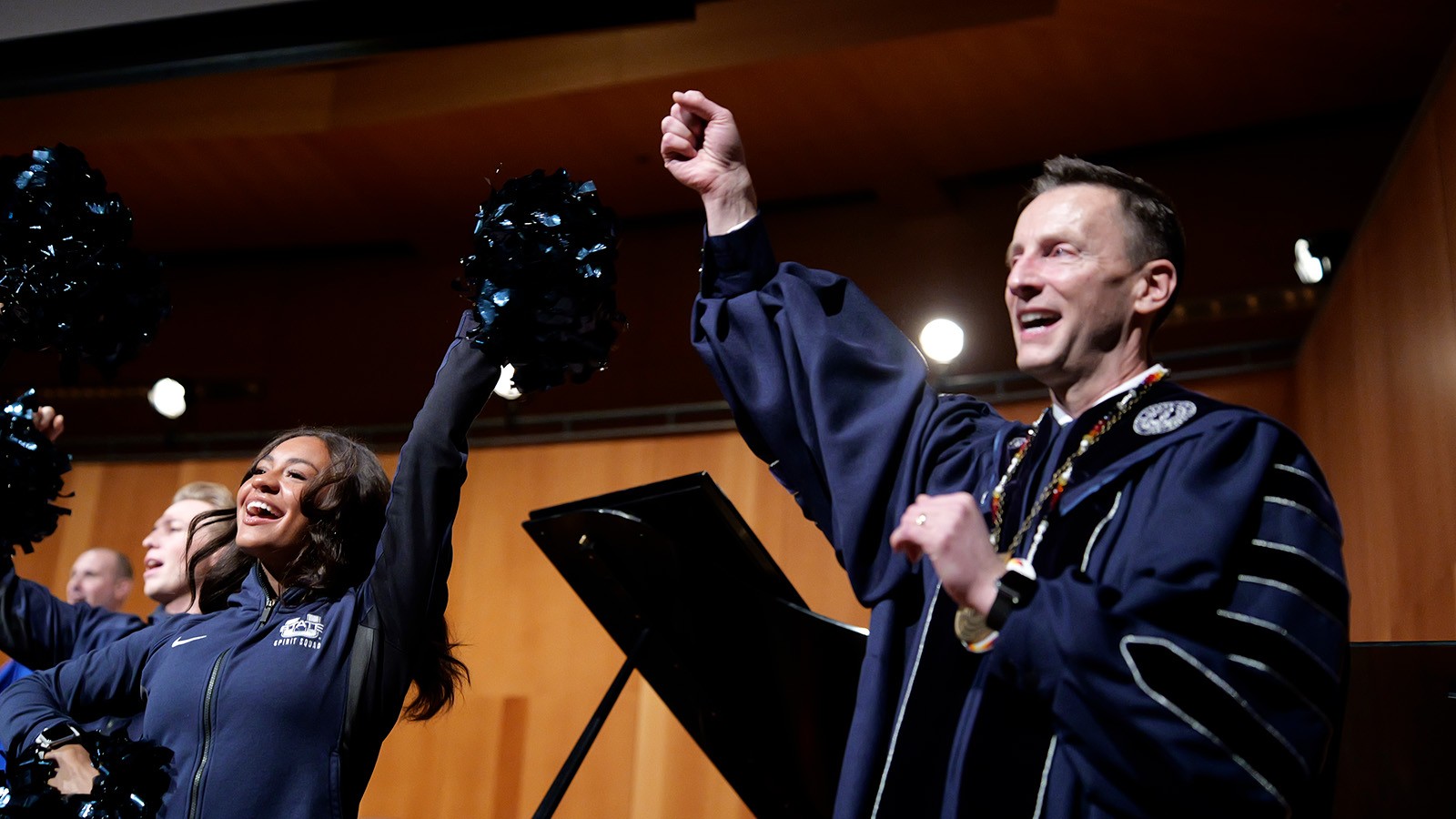 A man in regalia throws his hands in the air as part of the school fight song.