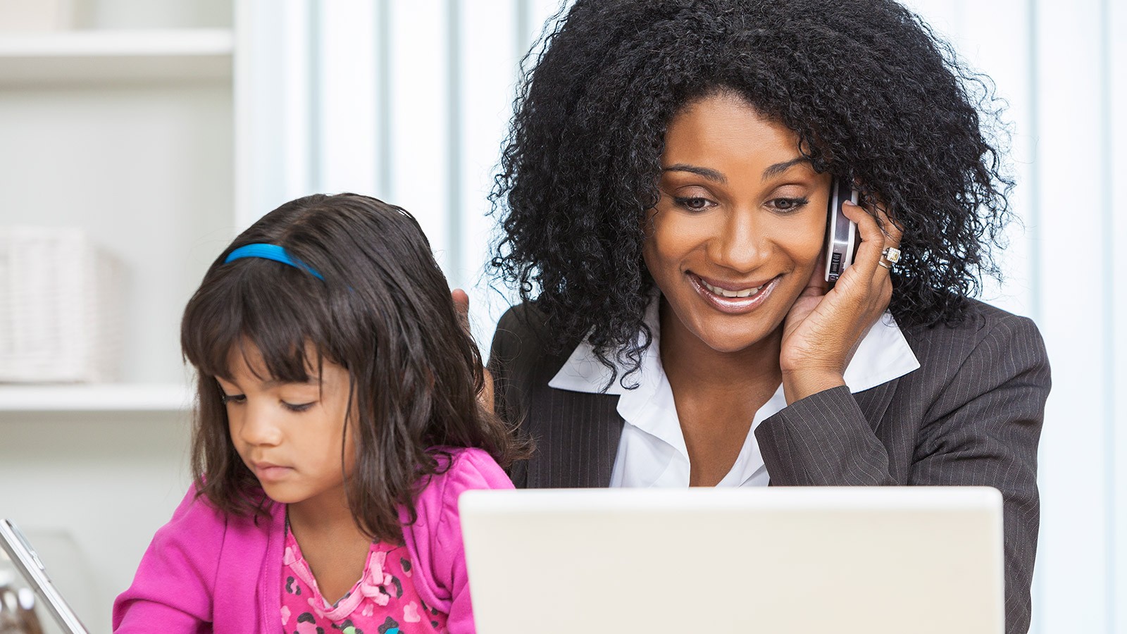 a mother holds her daughter on her lap while working on a laptop and talking on a phone.