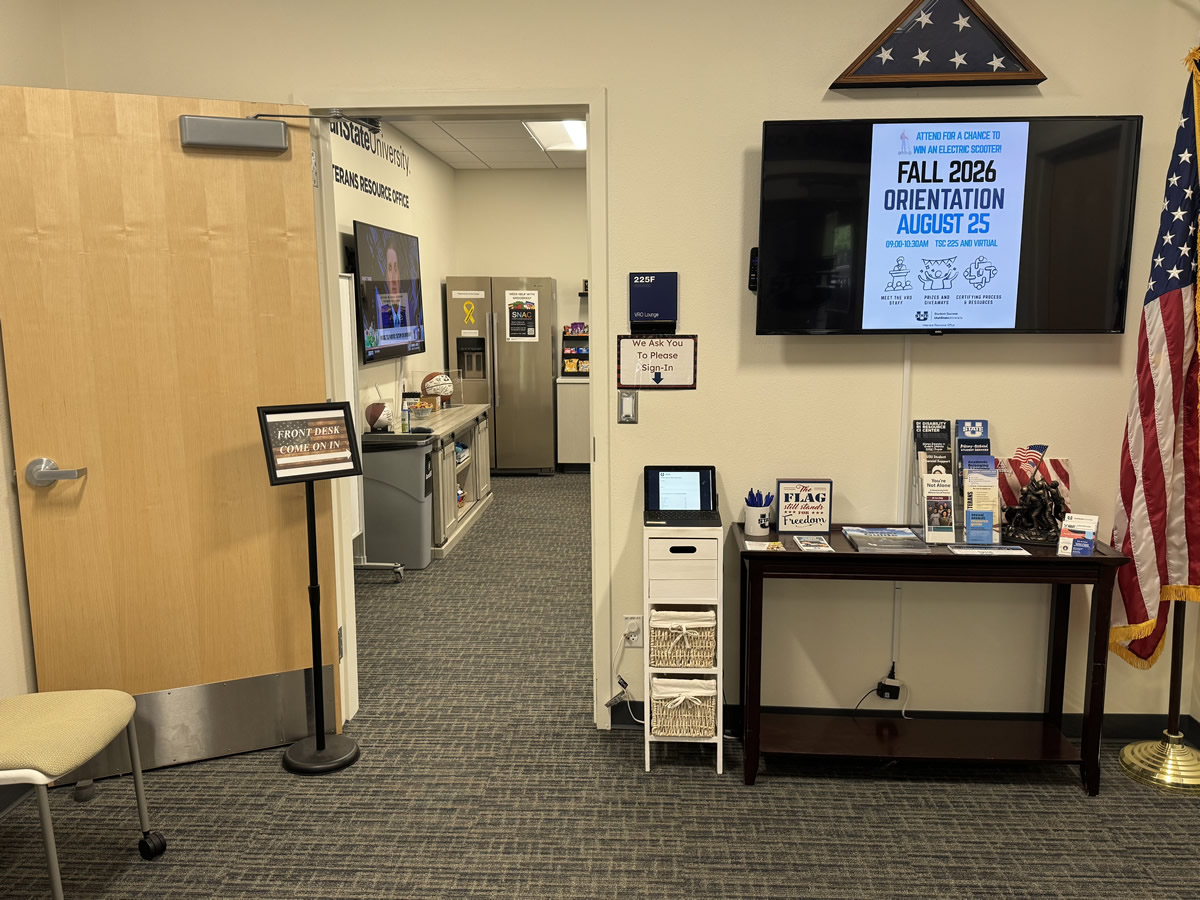 Welcome to the VRO, photo from the entrance door with a welcome to the VRO sign, a large TV screen that carries announcements, a sign-in registration computer, USA Flag on a stand and a tri-folded one in a case hanging on the wall, a small talbe with brochures and the entry way into the main lounge space.