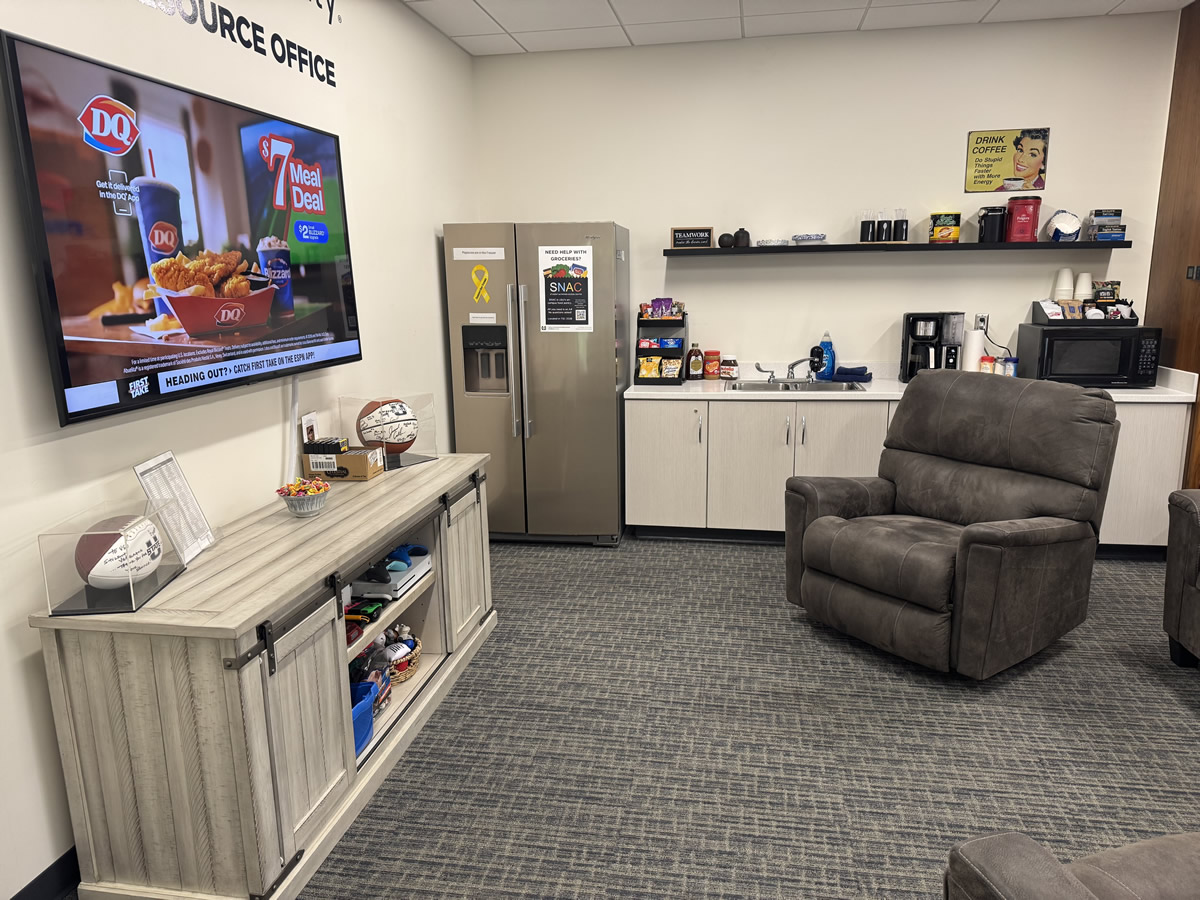 looking through the entry door glass of a credenza on the left against the wall with a large screen TV that plays movies and/or news through the day.  Past them on the back wall is a 2 door fridge, and a cabinet with a sink, coffee pot, microwave and rack with tea and snack selections, to the right is a large swivel recliner.