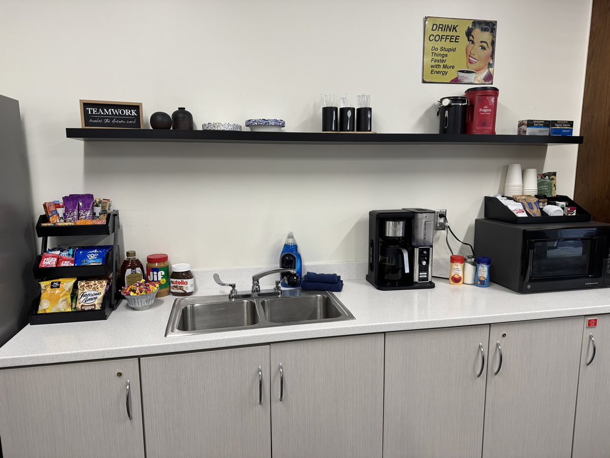 closer view of the counter top with the sink, coffee pot, microwave and rack with tea and snack selections. 